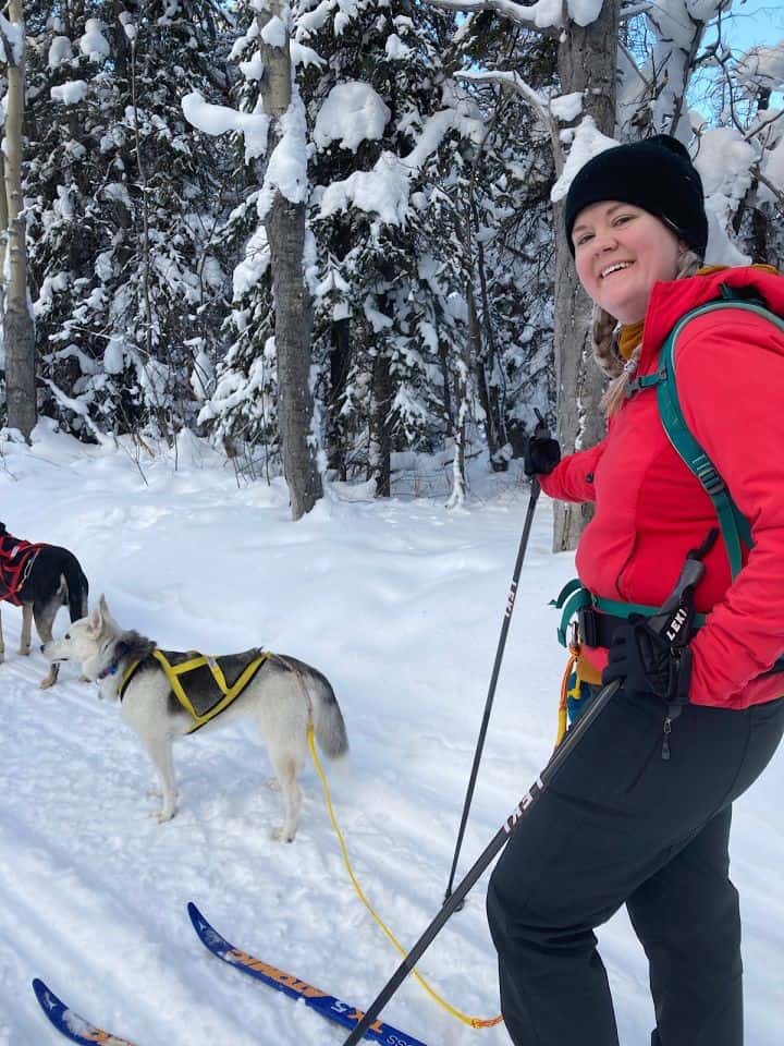 Woman in bright red jacket and harness stands with ski poles behind a husky in a yellow harness.