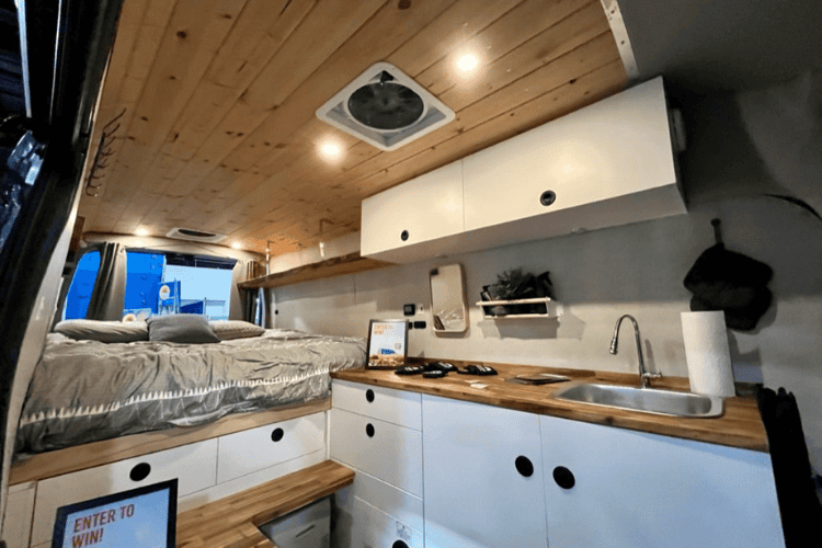 The interior of a camper van, equipped with a queen bed, cupboards, drawers, mirror, plants and even a kitchen sink.