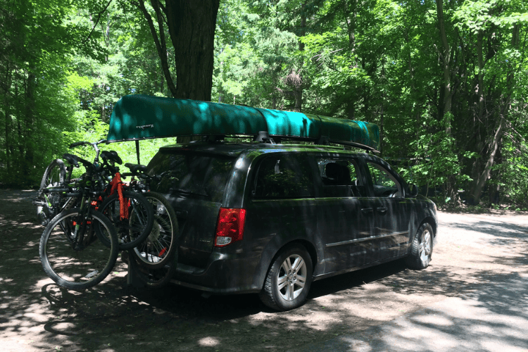 Dodge Grand Caravan parked in the forest, loaded with a green canoe and many bicycles.