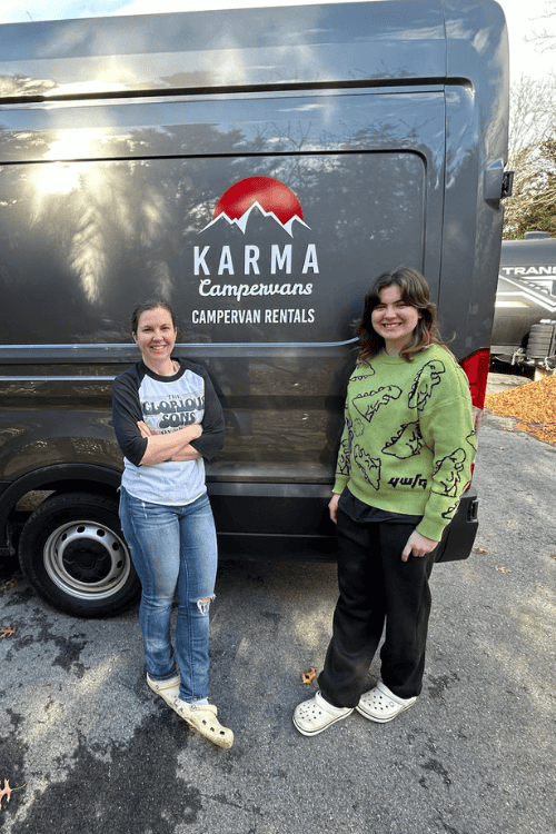 Two women pose, smiling, outside a camper van with the name 