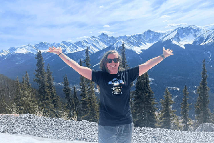 Woman stands with arms raised in front of many snow covered mountain peaks in the Canadian Rocky Mountains.