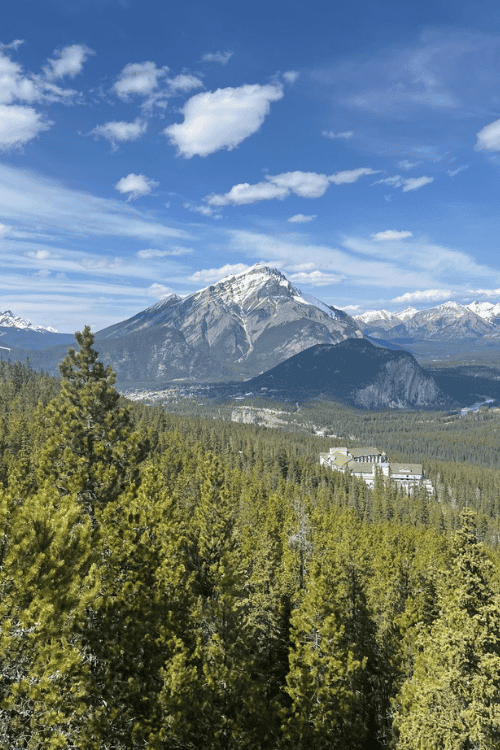 Snowy mountain peaks over a lush green forest, with blue sky and white clouds Banff, Alberta.