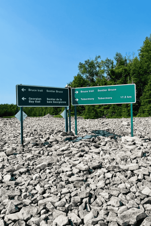 Green Parks Canada trail signs stand amongst the rocks on a rock-strewn beach along the Bruce Trail.
