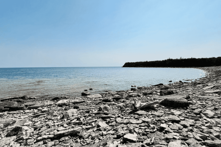 A grey rock strewn beach with blue waters on a beautiful blue sky day.