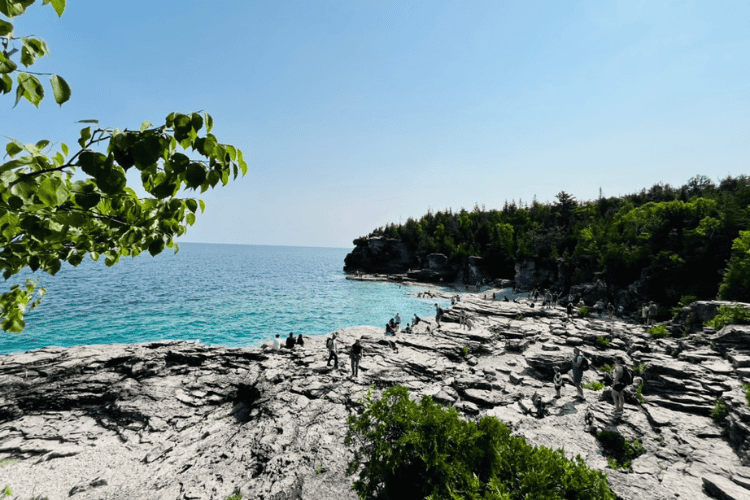 Smooth, grey, limestone beach of Indian Head Cove, bright aqua-blue waters of Georgian Bay and lush green forests of Bruce Peninsula National Park on a cloudless blue sky day.