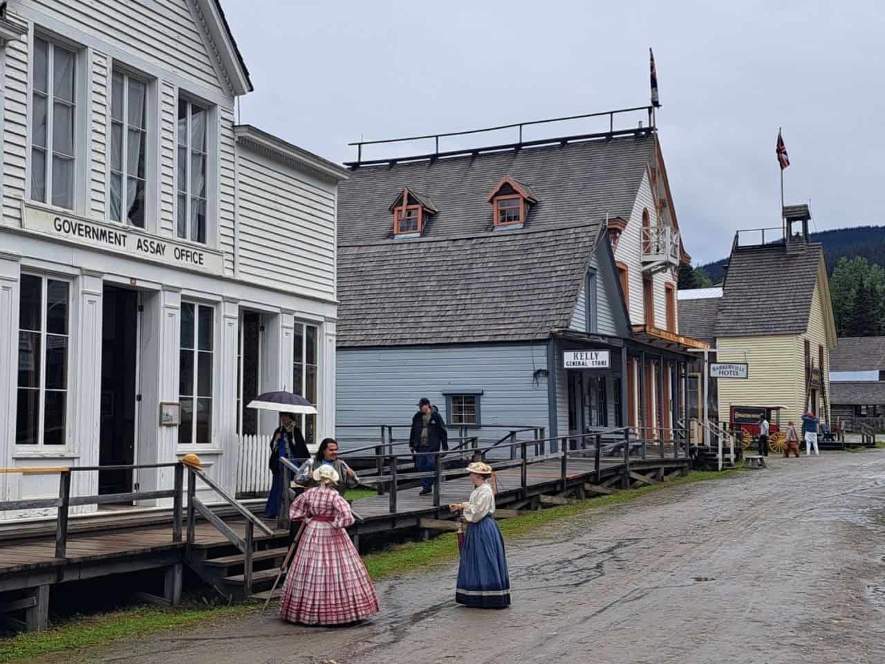 Barkerville Historic Town and Park  a muddy mainstreet in the rain. People dressed in period clothing stop to chat outside a store during the gold rush era.