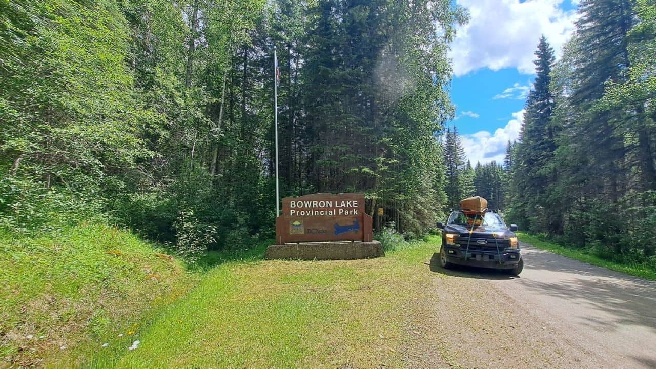 A black truck with a yellow canoe strapped to its rough stops next to the wooden Bowron Lake Provincial Park welcome sign.