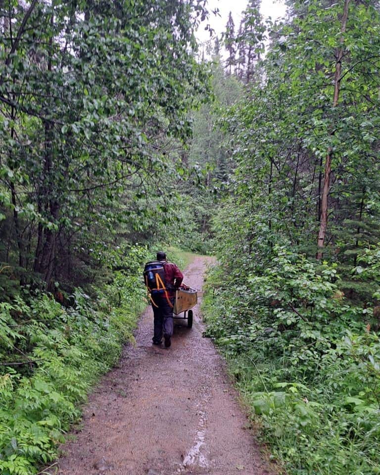 A man pushes a loaded canoe and carries a barrell on his back in the rain, along the registration ro Kibbee Lake portage on the Bowron Lakes Canoe Circuit