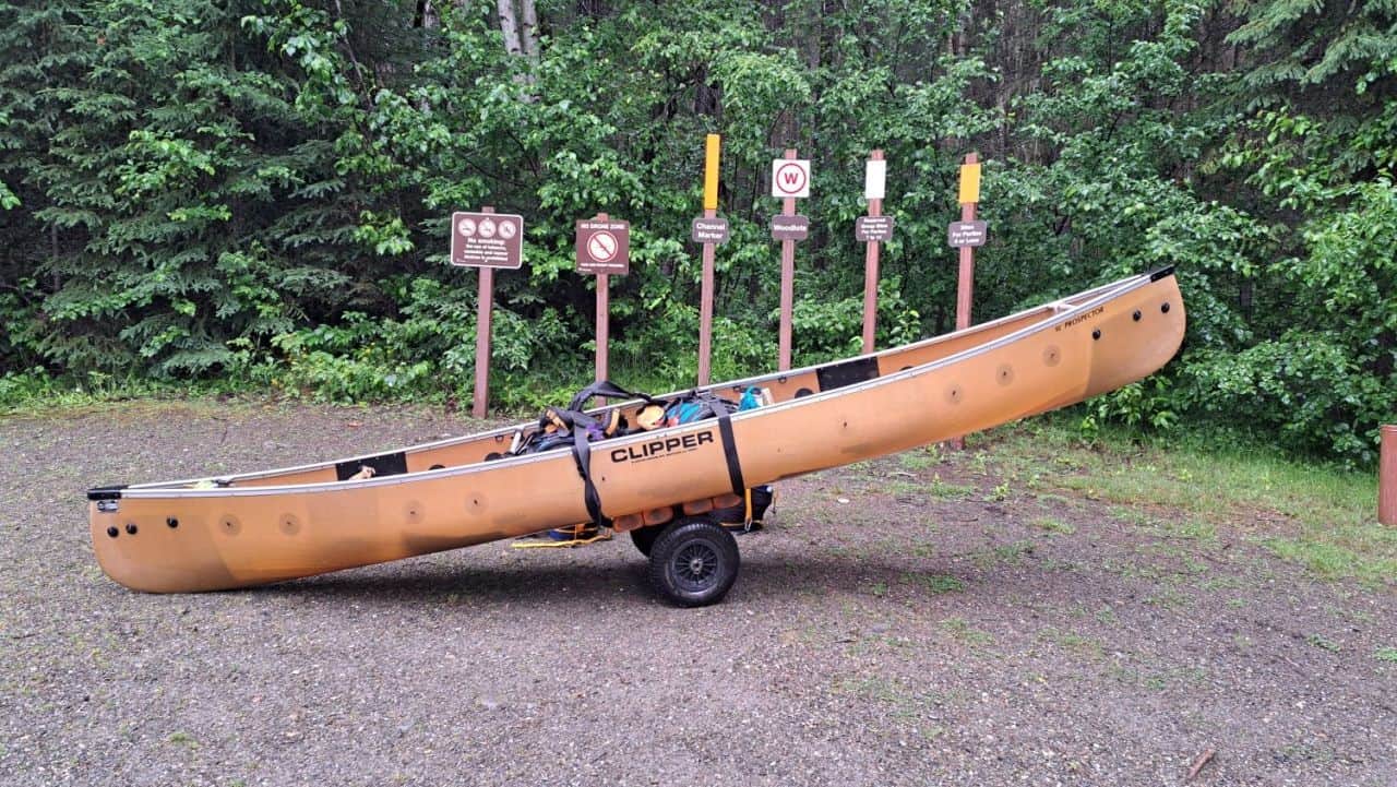 A canoe loaded onto an expedition canoe cart ready to start the first portage of the Bowron Lake Canoe Circuit in BC. Behind the canoe is a stand of six markers and signs for canoeists to watch for along the backcountry circuit.