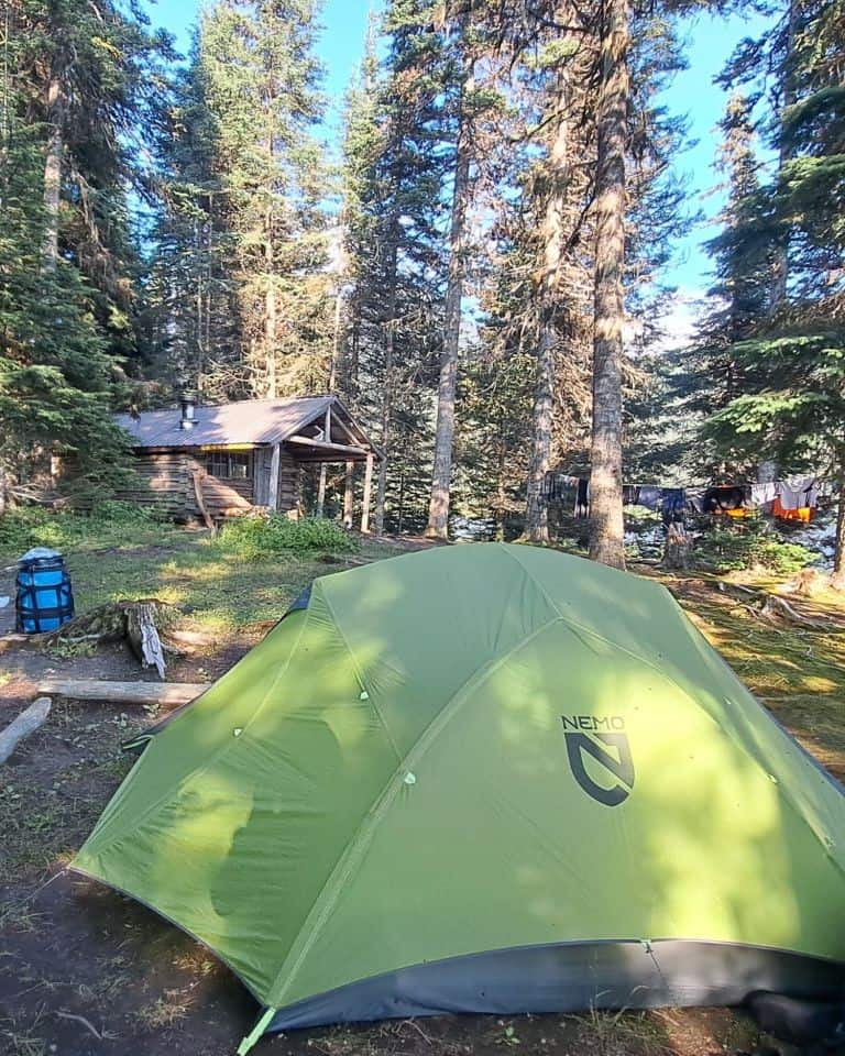 Campsite 8 on Indianlake on the Bowron Lakes Canoe Circuit in British Columbia, Canada shows a green Nemo tent in the foreground with a small historic log cabin in the background.