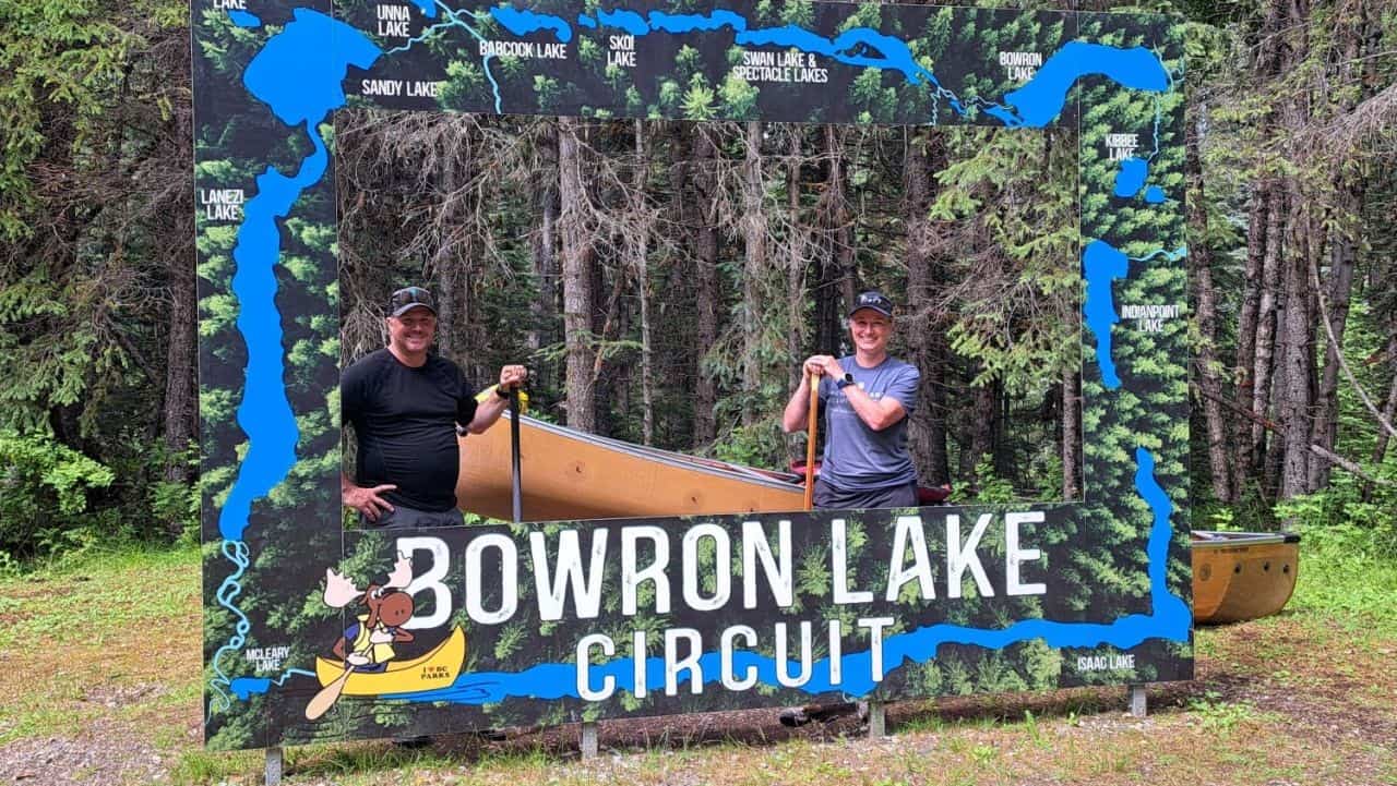 A couple poses with their yellow canoe while holding paddles. They are framed by the Bowron Lake Canoe Circuit map selfie station.