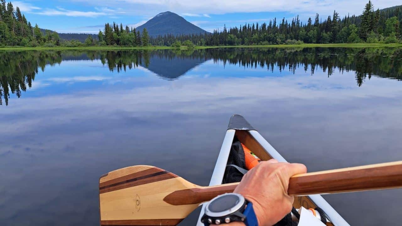 A lady holding a wooden paddle in the bow of the canoe takes a stroke on the calm waters of the westside of the Bowron Lakes Canoe Circuit. Ahead of her a pyramid shaped green mountain is reflected in the calm waters.