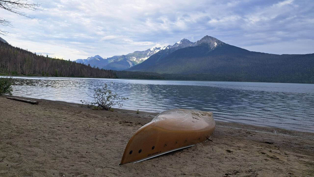 A yellow Tripper canoe is unpacked and flipped over for the night on a sandy beach on the aptly names Sandy Lake on Bowron Lakes. Across the lake's calm waters is a view of rugged snow capped mountains