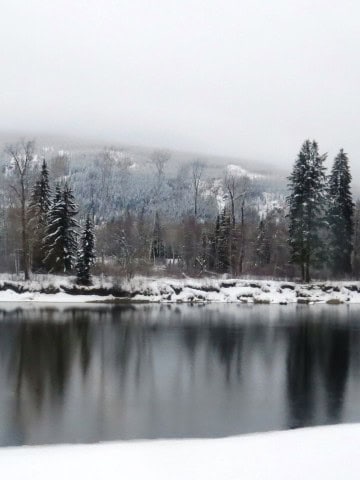 A still mountain lake near Jasper reflects snow-laden trees and rugged peaks, showcasing the serene beauty of Canada’s winter wilderness along VIA Rail’s The Canadian route through the Rockies.