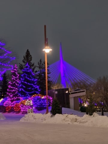 Outdoor holiday lights glow across The Forks in Winnipeg, reflecting off snow-covered walkways and frozen riverbanks during a winter stop on VIA Rail’s The Canadian transcontinental train journey.