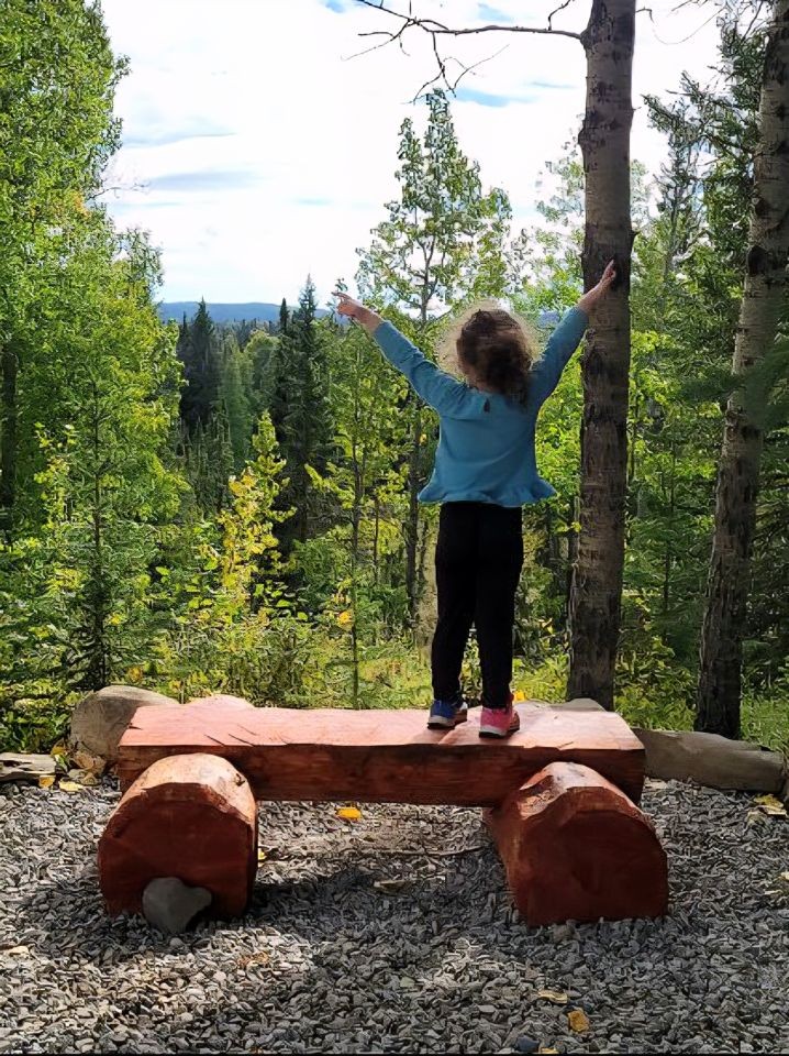 A child standing on a wooden bench with arms raised, surrounded by forest at Bearberry Cabins near Sundre, Alberta, Canada