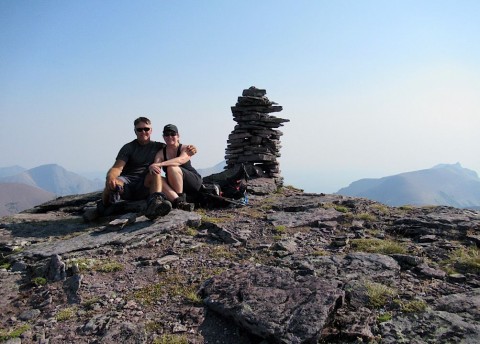 Man and woman sitting beside a large cairn on a mountain summit.