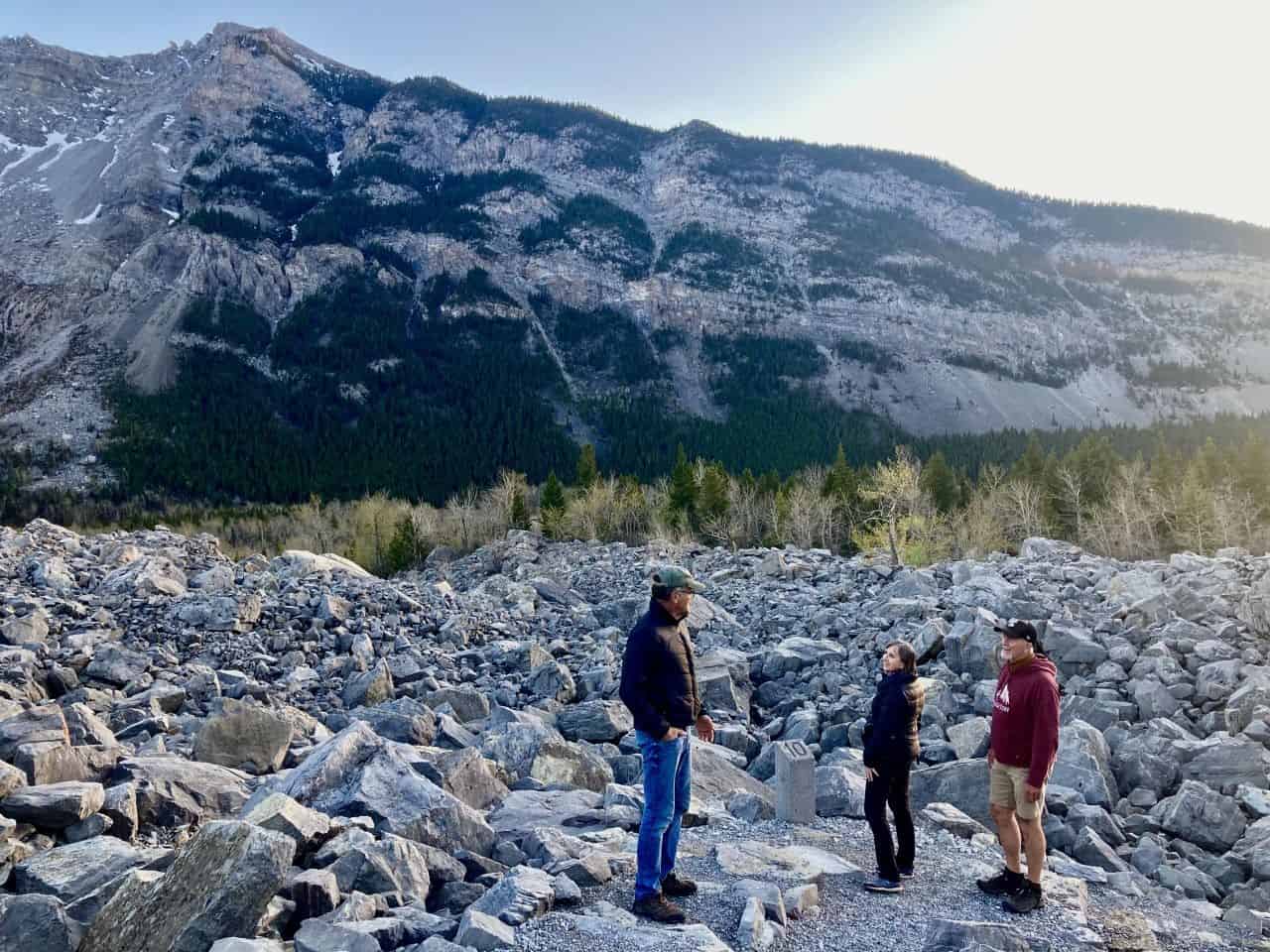 Two men and a woman standing among large boulders with mountain in background at Frank Slide.